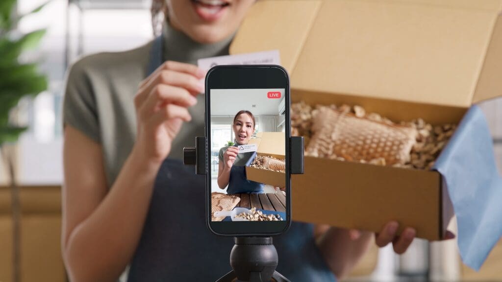 YouTube brand partnership deals: A content creator shares a product, holding a brown box with eco-friendly packing materials. Background is blurred but a smartphone in portrait mode on a tripod is focused on the scene.